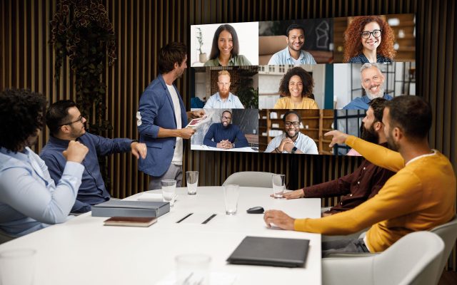 People working around a table, looking at a large LED screen broadcasting a meeting live